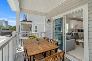 Eine Holzterrasse mit Tisch und Stühlen, eine Schiebetür zu einem Wohnbereich mit Sofa und Couchtisch sowie Blick auf die Stadtlandschaft.