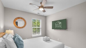 Un dormitorio con cama, ventilador de techo, ventana, espejo montado en la pared y un cartel enmarcado.