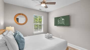 Une chambre à coucher comprenant un lit, un ventilateur de plafond, une fenêtre, un miroir fixé au mur et un panneau décoratif encadré.