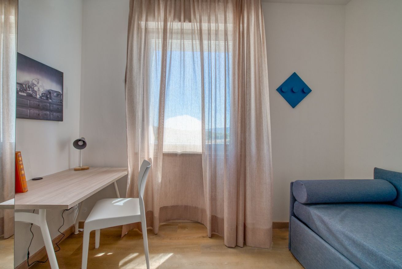 A room with a desk, chair, bed, and a window with sheer curtains.