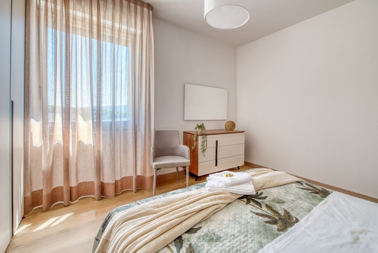 A bedroom with a bed, a chair, a dresser, and a window with sheer curtains.