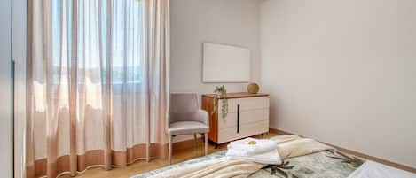 Un dormitorio con cama, una silla, un aparador y una ventana con cortinas traslúcidas.