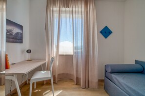 Een kamer met een bureau, stoel, bed en een raam met dunne gordijnen.