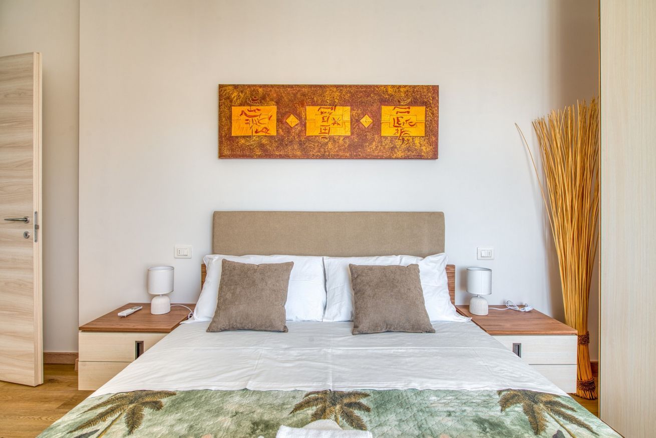 Une chambre d’hôtel comprenant un lit, deux tables de chevet, une petite table d’appoint, une œuvre d’art accrochée au mur et une grande plante décorative.