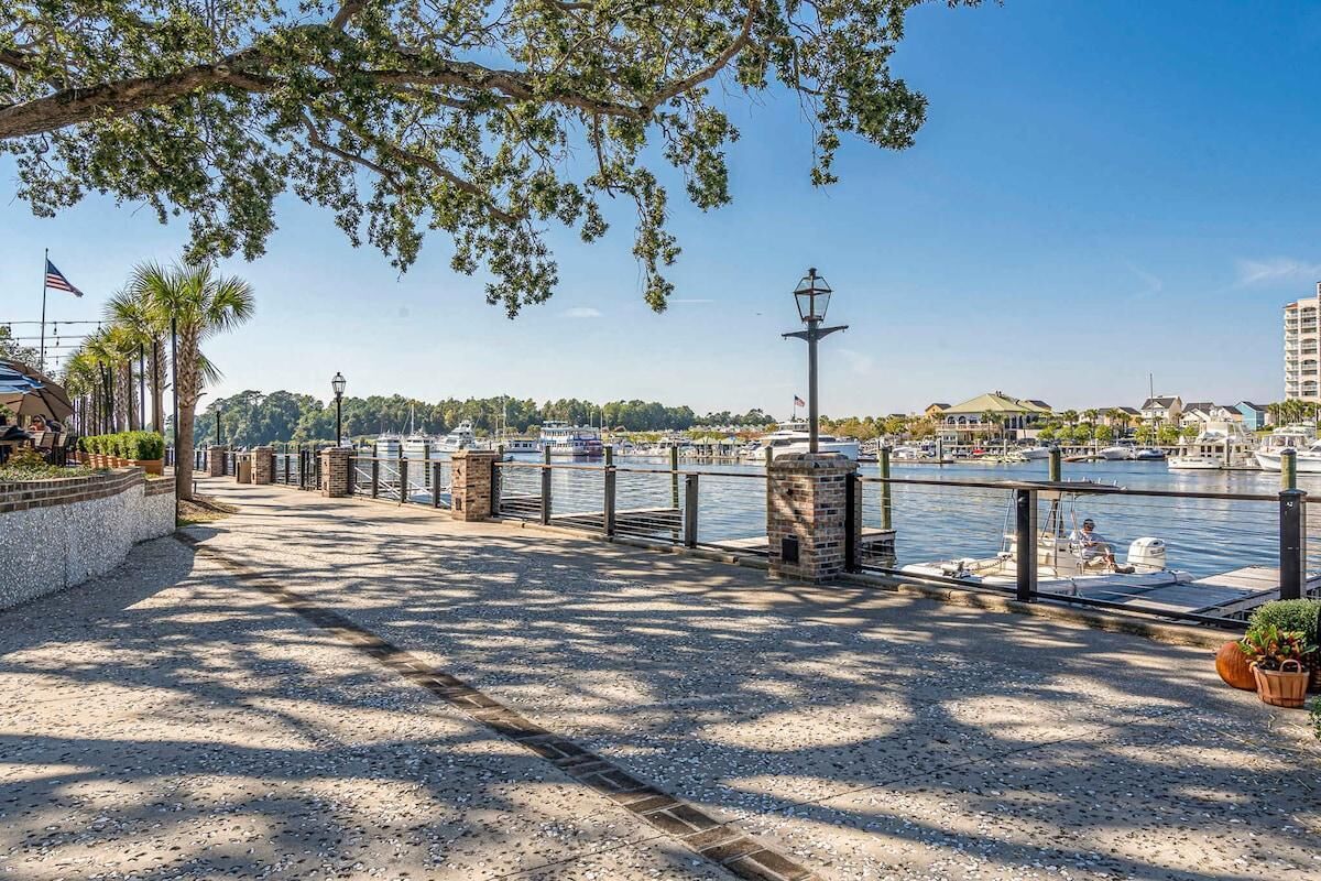 A waterfront promenade with a gravel path, a metal railing, and a view of boats and buildings across the water.