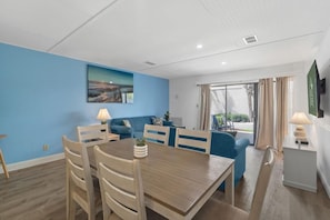 A modern living room with a dining area, featuring a blue wall, wooden furniture, and a large window with curtains. - Myrtle Dunes- Across From Beach, Pet Friendly (Myrtle Beach)