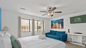 Quarto de hotel com cama, sofá azul, escrivaninha e ventoinha no teto.