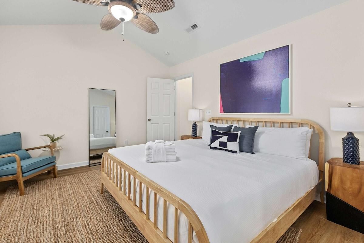 A bedroom with a wooden bed, a blue chair, a ceiling fan, and a large abstract painting on the wall.