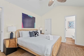 Un dormitorio con cama de madera, ropa de cama blanca, ventilador de techo y un cuadro abstracto grande en la pared.