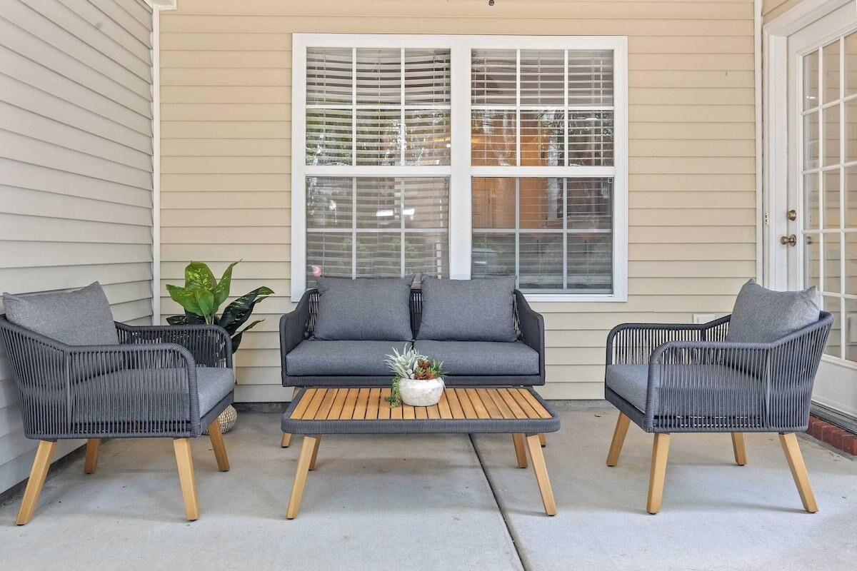 Une terrasse avec trois fauteuils en osier et un canapé, une table basse en bois agrémentée d’une plante, et une grande fenêtre avec des stores.
