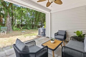 Une terrasse couverte, meublée de meubles en osier, dotée d’un ventilateur de plafond et offrant une vue sur un jardin avec des arbres et un barbecue.