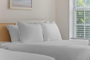 Un dormitorio con cama, ropa de cama blanca, cabecera y ventana con persianas.