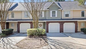 A two-story residential building with a brick facade, two attached garages, and a front yard with a small tree and shrubbery. - Boho Ferry- Mins From Credit One- Pet Friendly (Charleston)