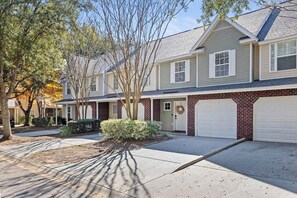 A two-story residential building with a brick facade, white garage doors, and a front yard with trees and shrubs. - Boho Ferry- Mins From Credit One- Pet Friendly (Charleston)