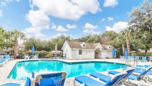 A swimming pool with blue lounge chairs and umbrellas, surrounded by trees and a building with a sloped roof. - Boho Ferry- Mins From Credit One- Pet Friendly (Charleston)