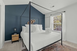 Een slaapkamer met een hemelbed, nachtkastje, raam met gordijnen en een decoratief wandobject.