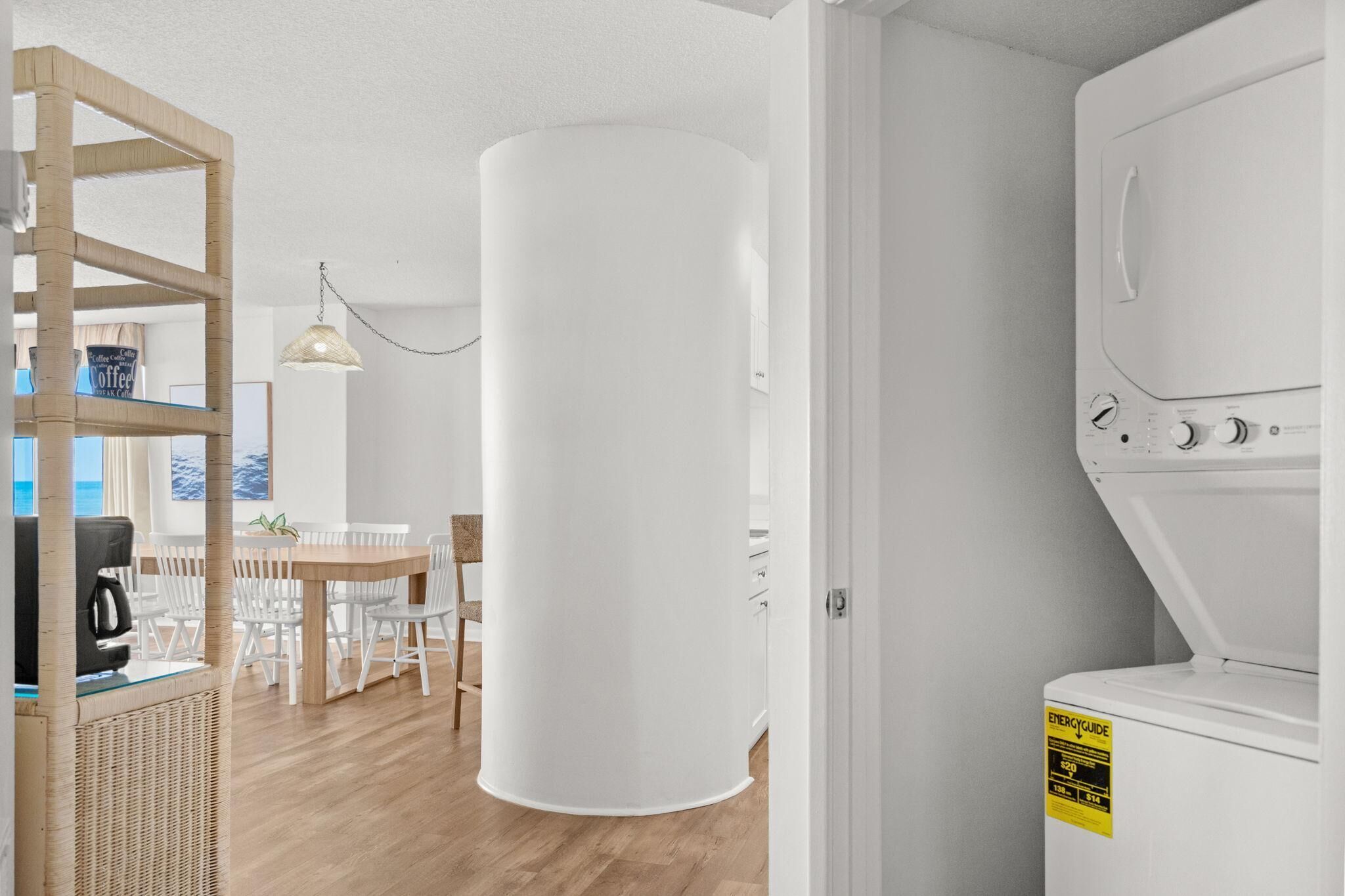 A room with a large cylindrical white object, a white washing machine, a dining area with a table and chairs, and a wicker shelf.