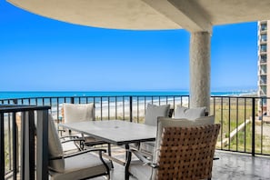 Een balkon met een tafel en stoelen, uitzicht op het strand en een gebouw op de achtergrond.