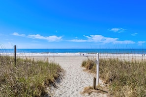 Een zandpad langs het strand dat uitkomt bij de oceaan, met een heldere lucht.