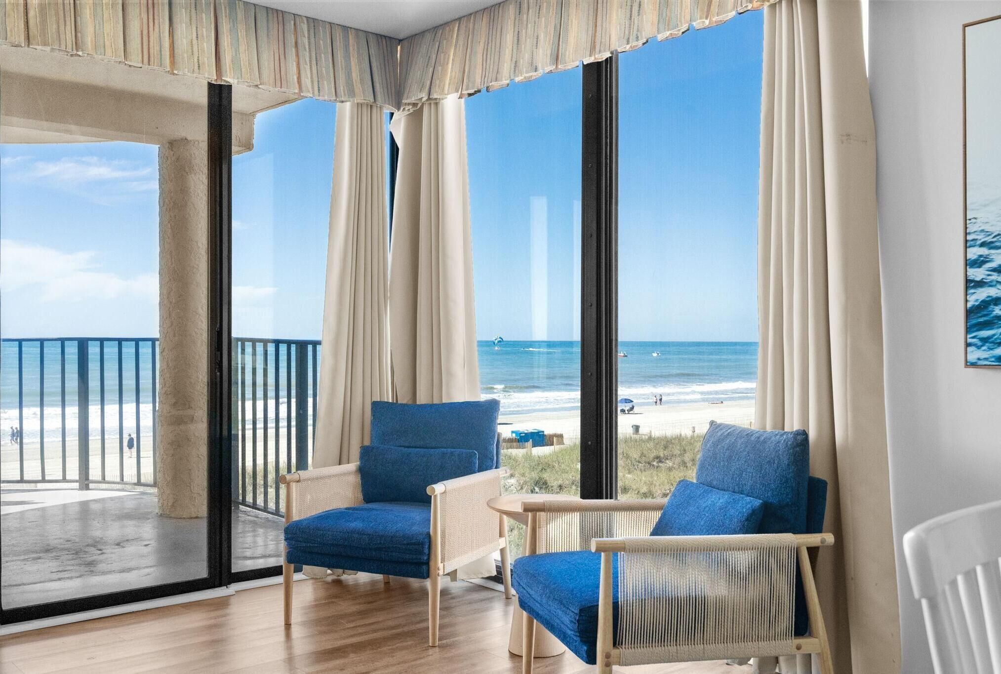 A room with large windows, a balcony, two blue chairs, and a view of the beach.