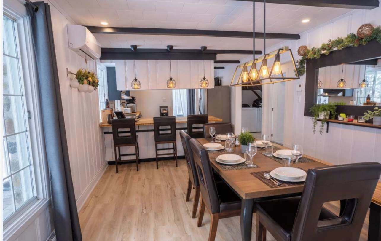 Un coin repas avec une table en bois dressée pour quatre personnes, une cuisine en arrière-plan et des luminaires suspendus au-dessus.