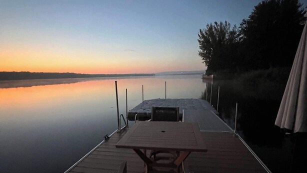 A wooden dock extending into calm water with a table and chairs, surrounded by trees.