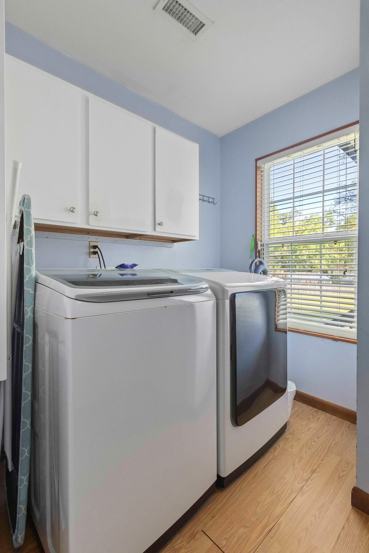 A laundry room with a washing machine and dryer, wooden flooring, a window with blinds, and white cabinets.