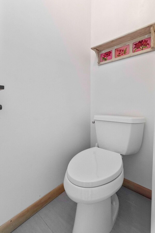 A white toilet with a wooden baseboard and a wall-mounted shelf with floral decorations. - The Endless Summer-hot Tub-7 Beds (North Charleston)