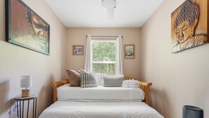 Une chambre à coucher avec un lit, une fenêtre avec des stores, une petite table avec une lampe et des œuvres d’art accrochées aux murs.