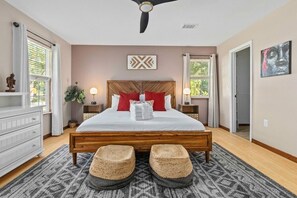 Ein Schlafzimmer mit einem hölzernen Bett, roten und weißen Kissen, einem Deckenventilator und einem großen Fenster mit Vorhängen.