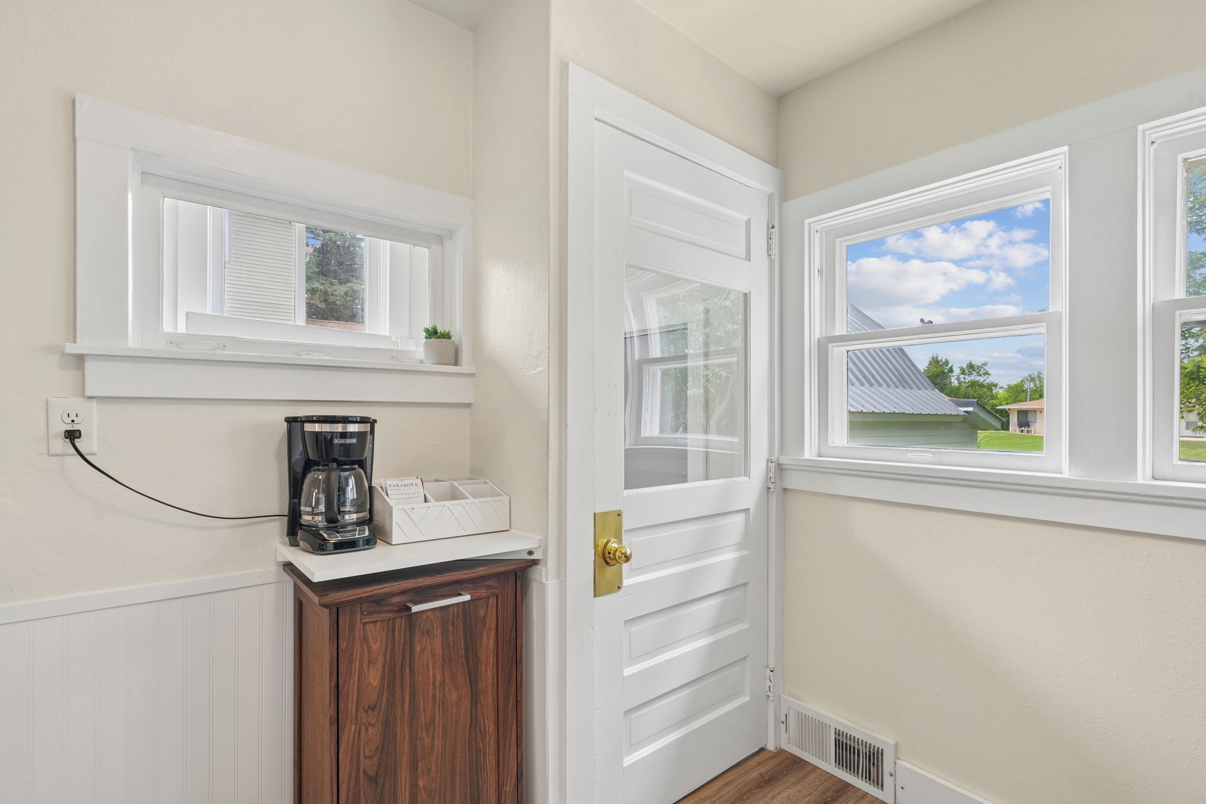A room with a white door, a window with a view of a house, a coffee maker on a shelf, and a power outlet.
