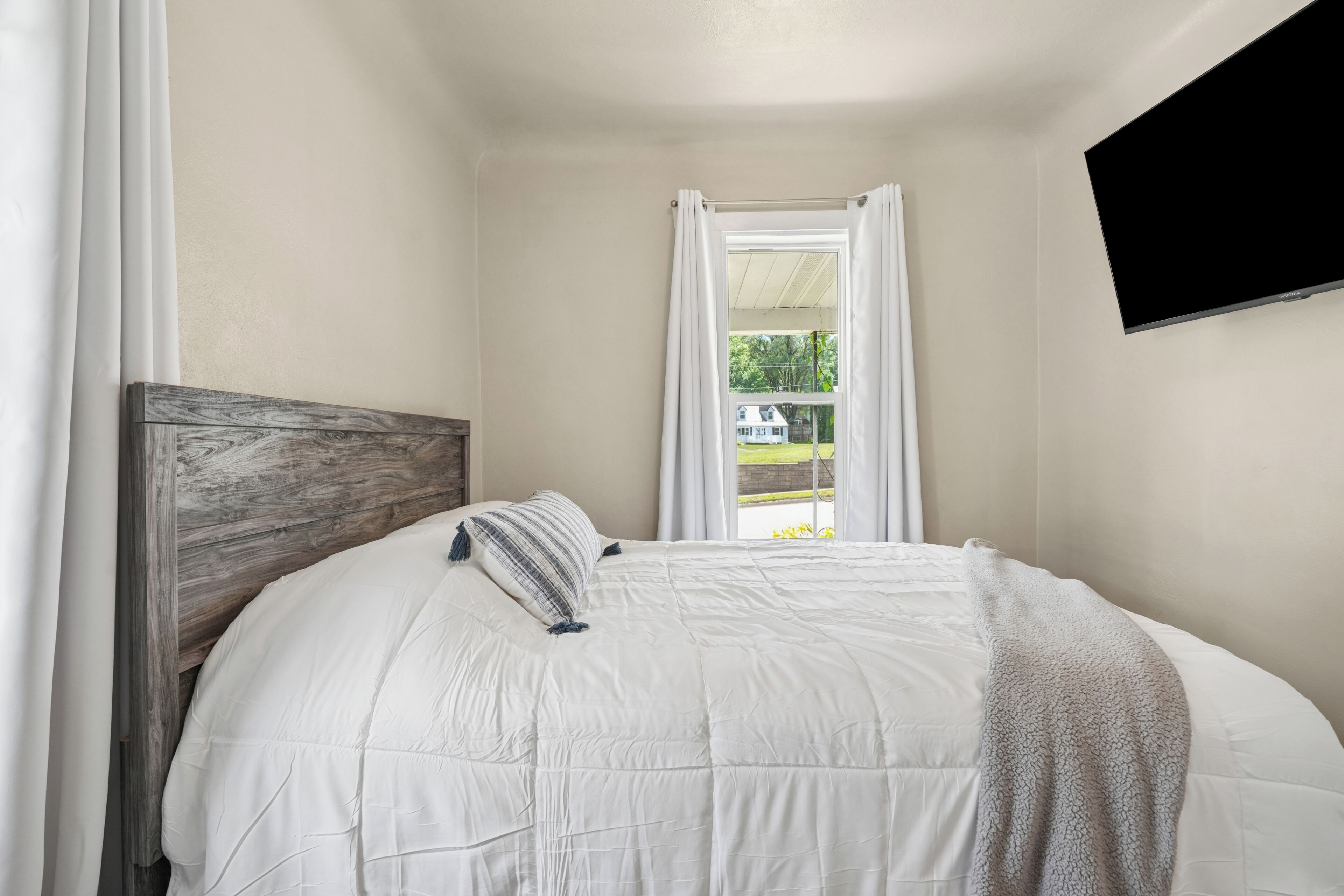 Un dormitorio con cabecera de madera, cubrecama blanco, ventana con cortinas blancas y un televisor de pantalla plana montado en la pared.
