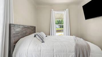 Un dormitorio con cabecera de madera, cubrecama blanco, ventana con cortinas blancas y un televisor de pantalla plana montado en la pared.