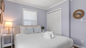 Een opgemaakt bed met witte lakens, een bedlampje en een raam met zonwering.