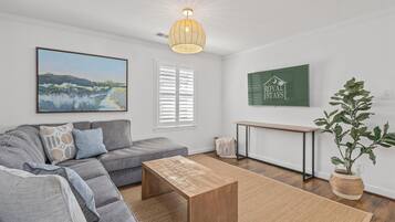 Een moderne woonkamer met een grijze bank, een houten bijzettafeltje, een schilderij, een groen bord en een potplant.