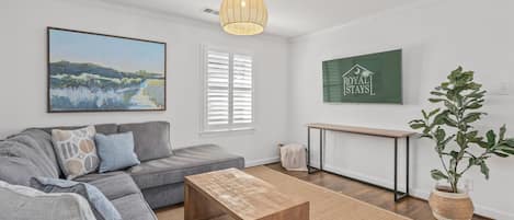 A modern living room with a grey sofa, a wooden coffee table, a painting, a green sign, and a potted plant.