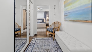 A hallway leading to a bedroom with a bed, a nightstand, and a lamp. There is a chair with a cushion and a bench with a cushion. A painting is on the wall, and a mirror is on the left. - Old Village Palmetto- Pet Friendly (Mount Pleasant)