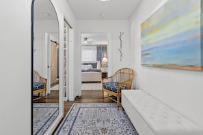 En gang der fører til et soveværelse med en seng, et natbord og en lampe. Der er en stol med pude og en bænk med pude. Et maleri hænger på væggen, og der er et spejl til venstre.