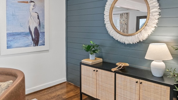 Een kamer met een donkerblauwe wand, een lijst met daarin een vogelafbeelding, een ronde spiegel met een witte verenomlijsting, een witte lamp en een rieten kast.