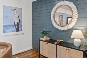 Um quarto com parede azul escura, um quadro de um pássaro, um espelho redondo com moldura branca de penas, um abajur branco e um armário de vime.