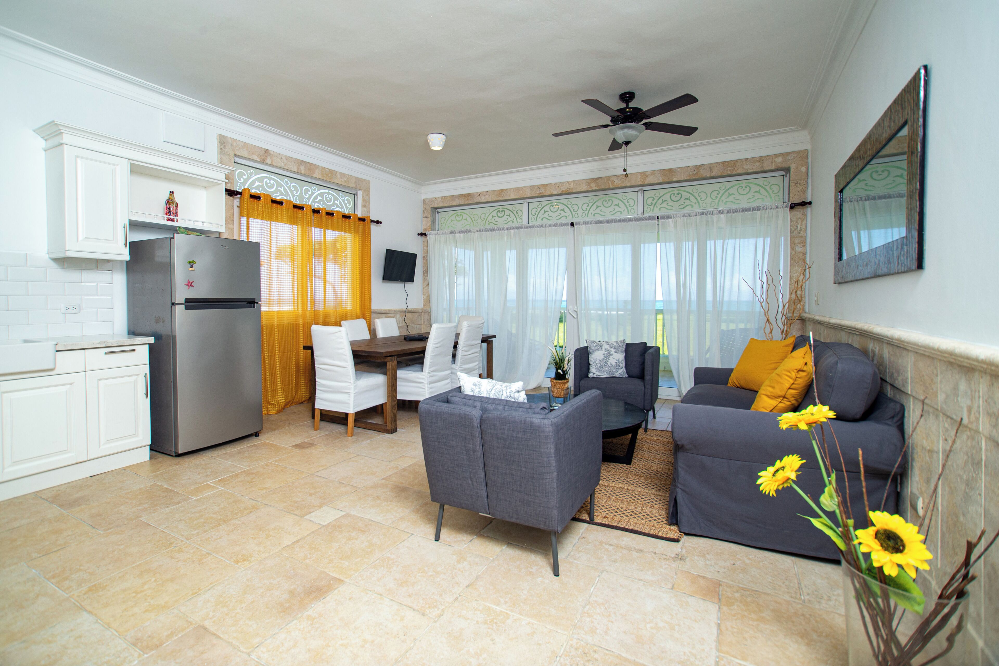 A modern kitchen with a refrigerator, dining table, and chairs. There is a living area with a sofa and a coffee table.