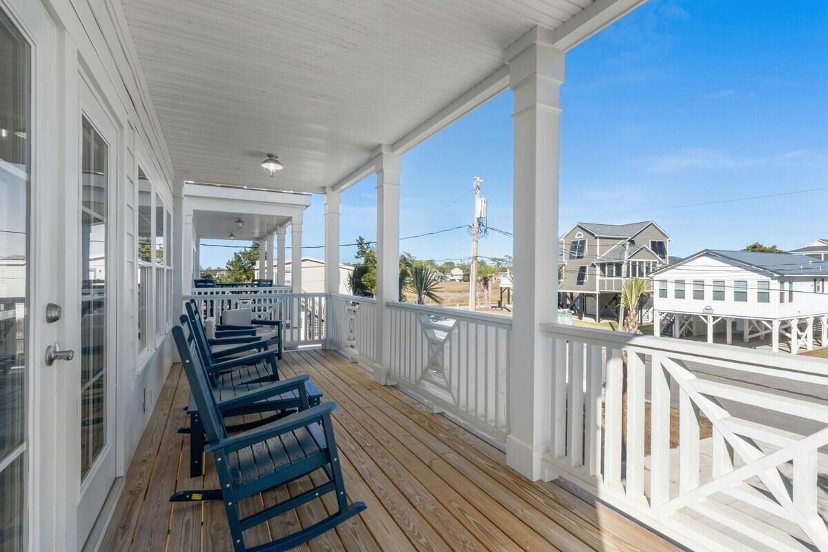 A covered porch with wooden flooring and white railings, overlooking a residential area with houses and a clear sky.