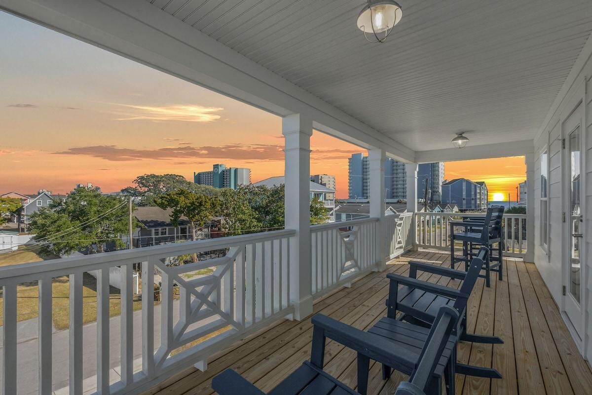 A covered outdoor area with wooden flooring and white railings, featuring outdoor seating and a view of the city skyline at sunset.