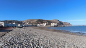 Una spiaggia di ciottoli con una passeggiata, edifici e una collina sullo sfondo.