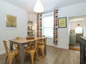 A dining area with a wooden table and chairs, a pendant light, a bookshelf, and a window with a patterned curtain. - Mona Lodge (Llandudno)