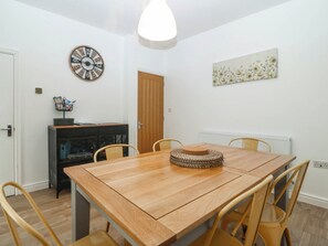 A dining area with a wooden table, four chairs, a decorative wall clock, a framed floral artwork, and a basket on a cabinet. - Mona Lodge (Llandudno)
