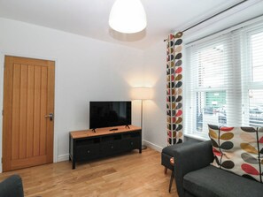 A living room with a television on a wooden stand, a floor lamp, a sofa, and a window with blinds. - Mona Lodge (Llandudno)