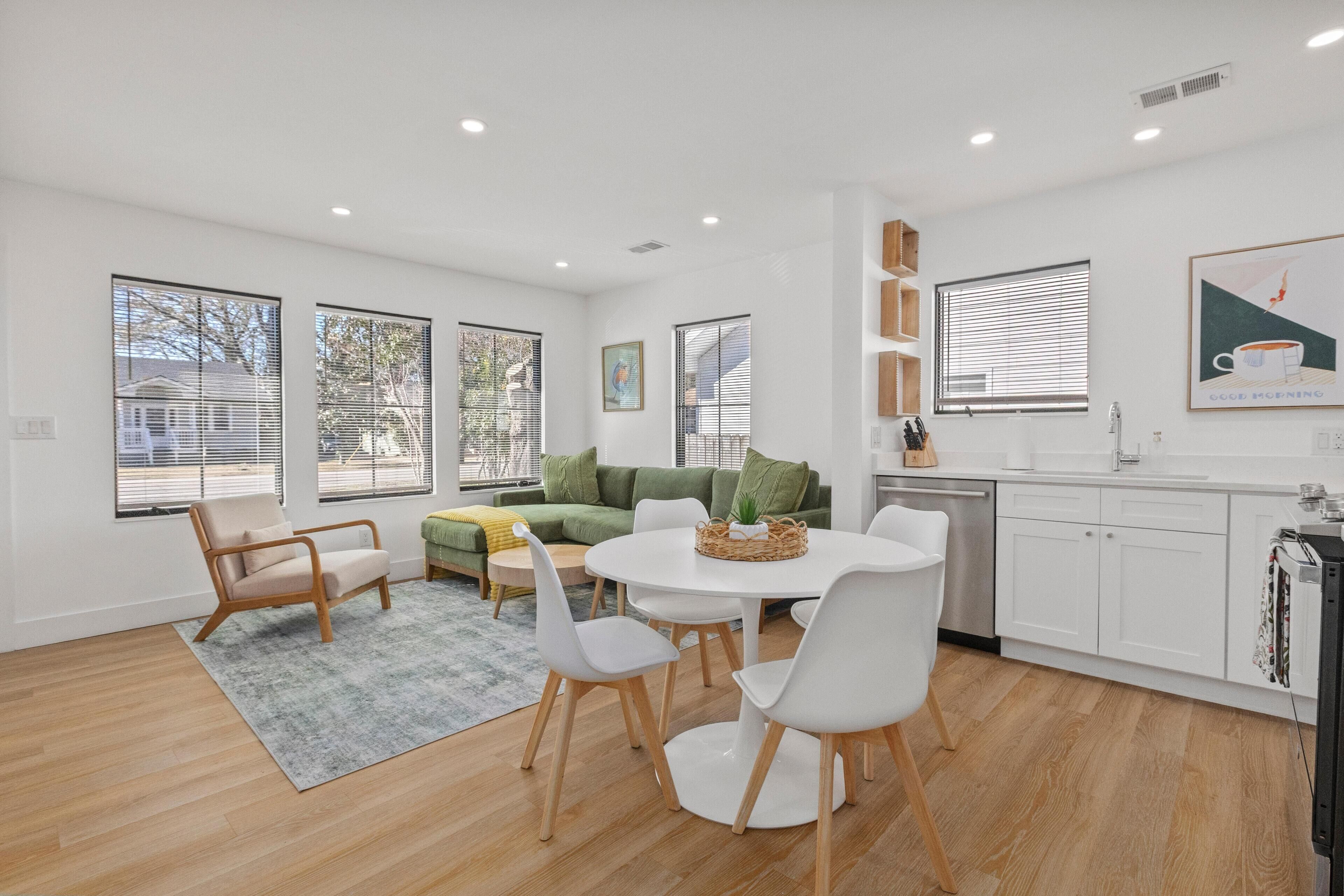 A modern, well-lit living room with a round dining table, white chairs, a green sofa, and a kitchen area with white cabinets and a sink.