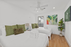 A hotel room with two beds, a ceiling fan, a window with blinds, a painting, and a potted plant.
