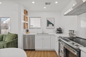 A modern kitchen with white cabinetry, a stainless steel dishwasher, a coffee machine, and a window with blinds.